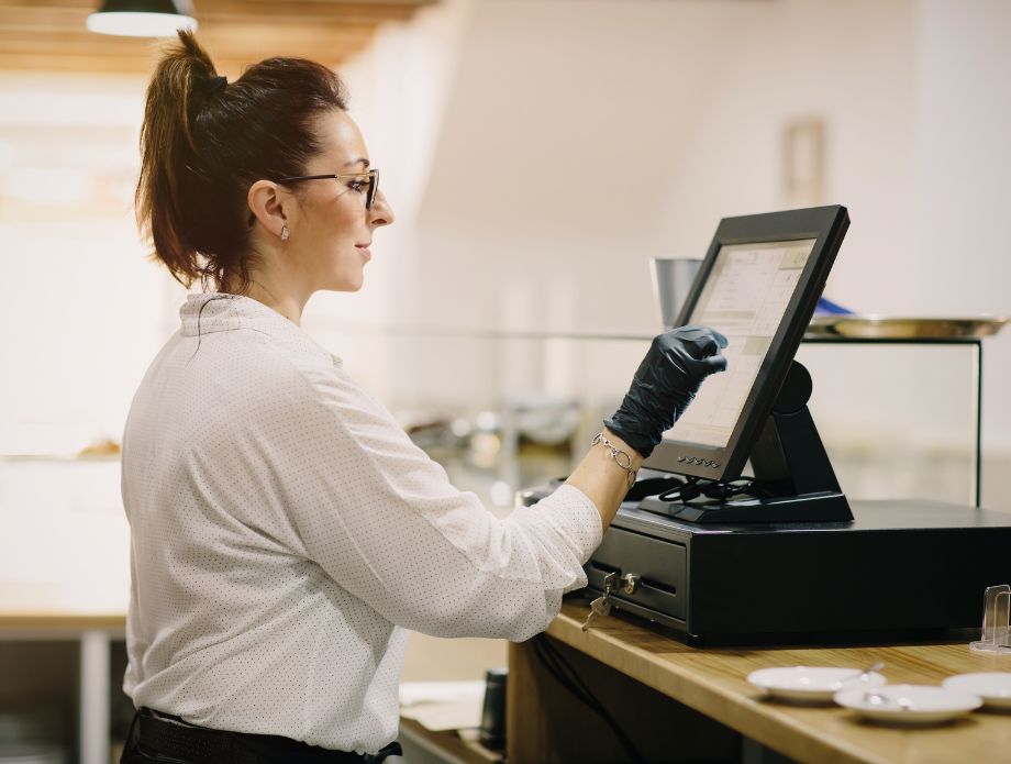 Una mujer con gafas y guantes negros operando una caja registradora electrónica en un restaurante.